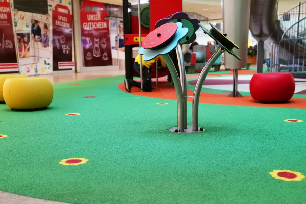 Ein interaktives Klangspielgerät in Blumenform mit drehbaren bunten Elementen steht auf einer grünen Spielfläche im Einkaufszentrum. Umgeben ist es von gepolsterten Sitzelementen und weiteren Spielstrukturen im Hintergrund.