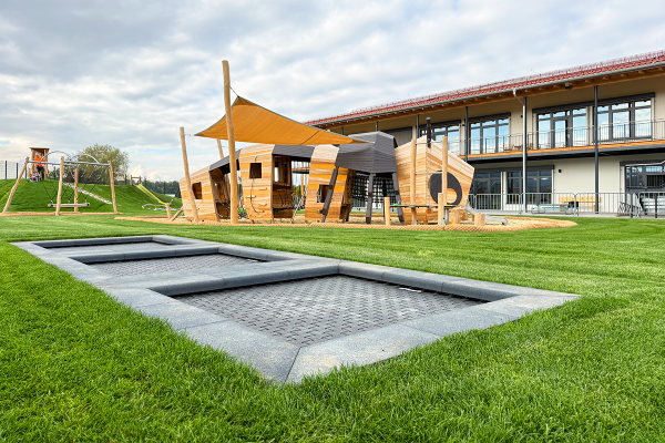 Moderner Kindergarten-Spielplatz mit großem Naturholz-Spielgerät in Bienenform, integriertem Sonnensegel, Bodentrampolin und weitläufigem Grünbereich vor einem Kindergarten-Neubau.
