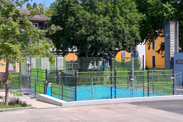 Multisportanlage mit zwei Basketballkörben, Ballfangzaun und blauem Kunststoffbelag, eingebettet in eine grüne Schulumgebung in Lörrach