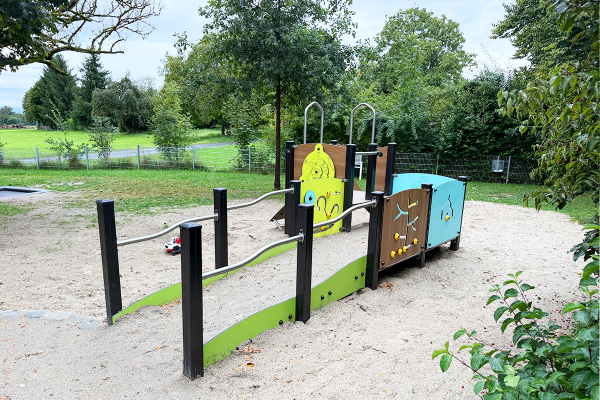 Barrierefreies Spielgerät mit Rampe, Handläufen und interaktiven Spielelementen auf Sandfläche in grüner Umgebung
