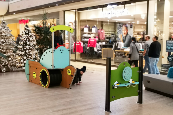 Ein maritim gestaltetes Spielgerät in Form eines Schiffes mit Kletterelementen, Kriechtunnel und Steuerrad steht auf hellem Boden in einem Einkaufszentrum. Ein Kleinkind krabbelt durch das Schiff, im Hintergrund sind Weihnachtsbäume und Schaufenster sichtbar.
