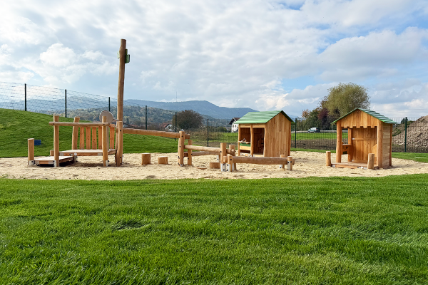 Spielbereich mit kleinen Holzhäuschen, Balancierbalken und Sitzhockern aus Naturholz auf einem Sandspielplatz mit Wiesenfläche und Bergpanorama im Hintergrund.