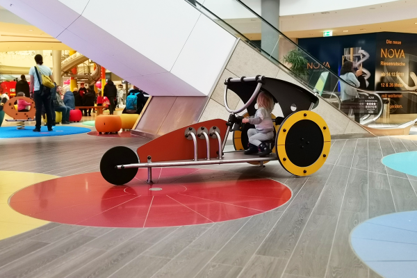 Ein Kleinkind sitzt in einem stilisierten Spielgerät in Form eines Dragsters mit großen gelben Rädern. Die Szene spielt sich auf einem farbigen Bodenelement vor einer Rolltreppe im Einkaufszentrum ab.