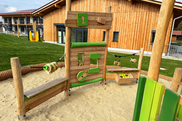 Detailansicht eines Holzspielbereichs mit grünen Akzenten, Spielwand, Fischspiel und integriertem Marktstand mit Holzäpfeln auf einem Sanduntergrund vor dem Gebäude des Kindergartens.