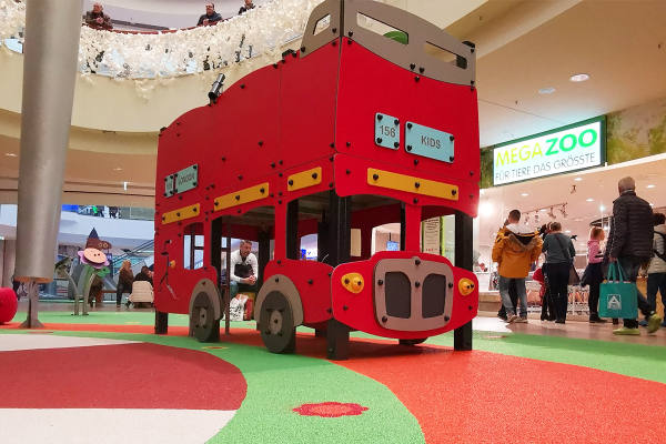 Ein großes, rot gestaltetes Spielgerät in Form eines Londoner Doppeldecker-Busses steht auf einer farbenfrohen Indoor-Spielfläche im Einkaufszentrum. Kinder können das Innere erkunden, während Erwachsene in der Nähe verweilen.