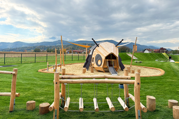 Spielgerät in Bienenform aus Naturholz mit Rutsche und Kletterelementen auf einem Spielplatz vor Bergkulisse, im Vordergrund eine wackelige Hängebrücke aus Holzstämmen.