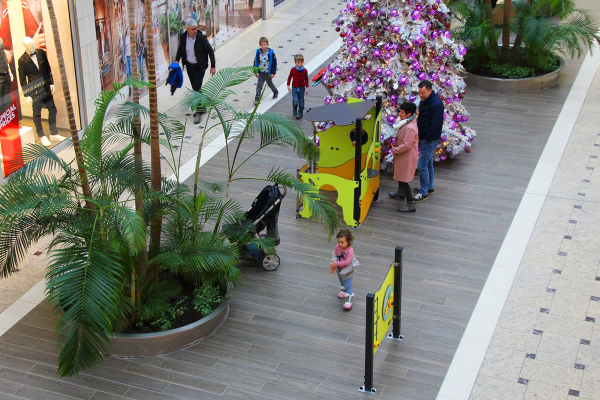 Ein grünes Spielhaus mit Kletter- und Spielelementen sowie eine separate Spielwand stehen auf einer großzügigen Flanierfläche im Einkaufszentrum. Kinder und Eltern interagieren, im Hintergrund ist ein geschmückter Weihnachtsbaum zu sehen.
