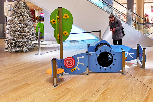 Ein niedriges, blaues Spielgerät mit Kriechtunnel und Spielanreizen für Kleinkinder steht auf hellem Boden vor einer Rolltreppe im Einkaufszentrum. Ein dekorativer Weihnachtsbaum und eine ältere Frau sind im Hintergrund zu sehen.