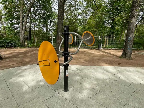Drehbares Outdoor-Fitnessgerät mit gelben Scheiben zur Mobilisation von Schultern und Armen, installiert auf einer barrierefreien Fläche im Park.