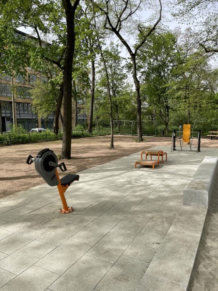 Barrierefreier Fitnessbereich im Mariannenpark Leipzig mit drei Outdoor-Trainingsgeräten auf gepflasterter Fläche, umgeben von Bäumen und Parkwegen.