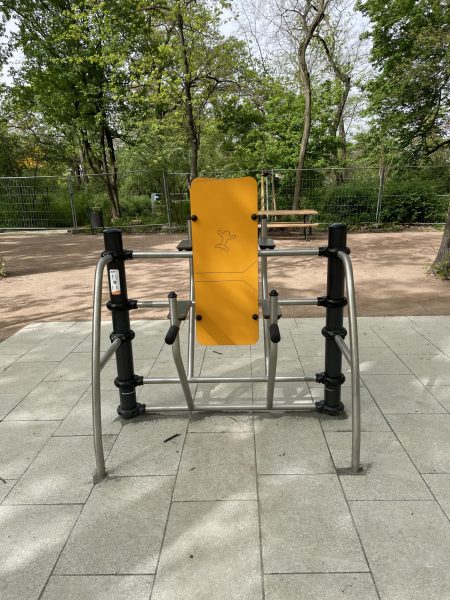 Gelbes Outdoor-Fitnessgerät mit Rückenlehne und Griffen auf einer barrierefreien Pflasterfläche im Mariannenpark Leipzig, umgeben von Bäumen.