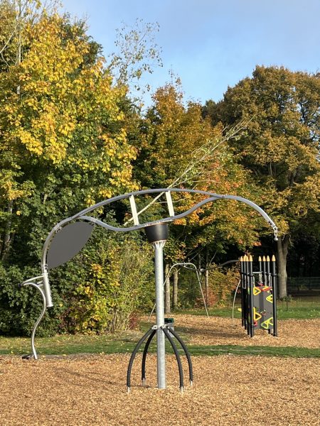 Das Outdoor-Fitnessgerät „Sky Surf“: ein großes, drehbares Balanceelement mit geschwungenen Haltearmen an einem zentralen Mast, installiert auf einem Holzhackschnitzelboden vor herbstlich gefärbten Bäumen.