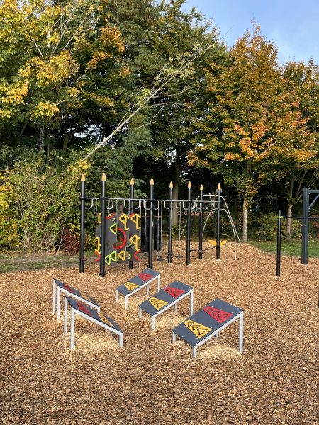 Outdoor-Trainingsbereich mit mehreren schrägen Sit-up-Bänken und einer Kletter- und Hangelstation im Hintergrund, umgeben von herbstlichen Bäumen und Holzhackschnitzelboden.