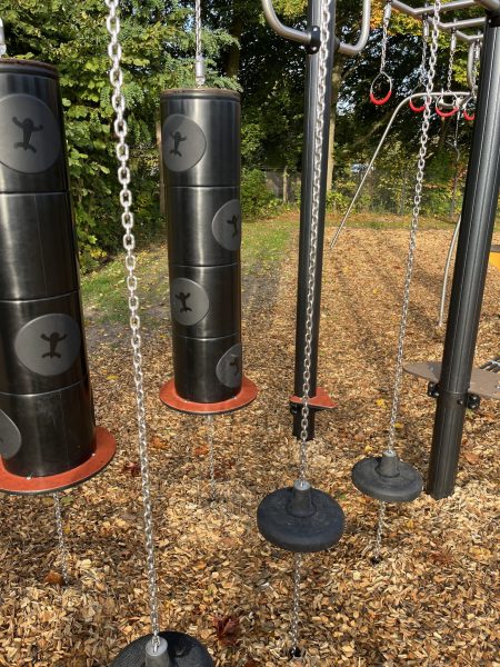Schwingende Balancier- und Trittmodule aus Gummi sowie hängende Trittzylinder an Ketten, die Teil eines beweglichen Parcours im Outdoor-Fitnessbereich sind; Holzhackschnitzelboden und herbstliche Bäume im Hintergrund.