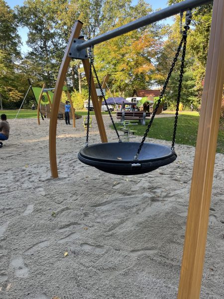 Nestschaukel auf Sandfläche im Eichpark Sottrum, im Hintergrund weitere Spielgeräte, spielende Kinder und Sitzgruppen unter Bäumen.