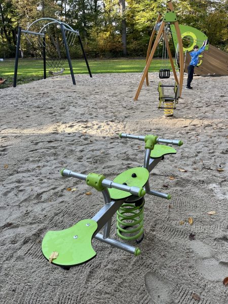 Spielgeräte auf Sandfläche im Eichpark Sottrum, darunter Wipptier, Kleinkindschaukel, Tampenschaukel und Spielschiff im Hintergrund, Kind spielt aktiv.