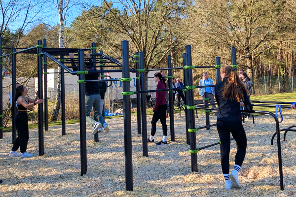 Menschen trainieren gemeinsam an einer Calisthenics-Anlage im Freien, umgeben von Bäumen und Zuschauer*innen.
