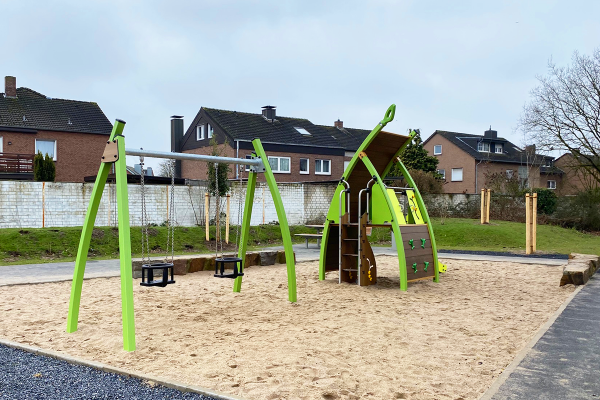 Spielbereich mit Kleinkindschaukeln und Kletterspielhaus auf sandigem Untergrund, umgeben von barrierefreien Wegen und begrüntem Wohnumfeld.