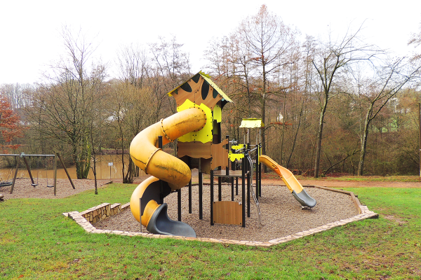 Großer Spielturm mit Röhrenrutsche, Kletterelementen und Wellenrutsche im Naturpark Kallenbach, umgeben von Bäumen und Teichlandschaft