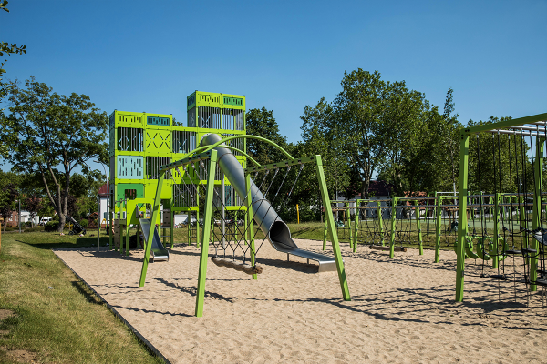 Grün gestaltete Klettertürme mit Edelstahlrutschen, Seilparcours und Balancierelementen auf einer Sandspielfläche im Zechenpark Kamp-Lintfort, eingebettet in eine parkähnliche Umgebung mit Bäumen.