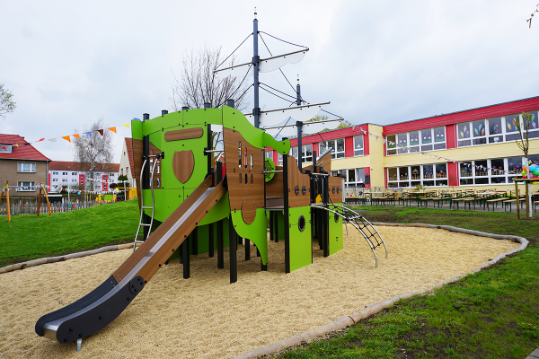 Großes grünes Spielgerät in Schiffsform mit Rutsche und Klettermöglichkeiten auf einem Kita-Spielplatz, im Hintergrund ein bunt geschmücktes Kita-Gebäude.