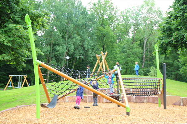Kinder spielen an einem großen Klettergerät mit Netz- und Balancierbereichen auf einem Spielplatz mit umliegender Wiese und Bäumen im Hintergrund.