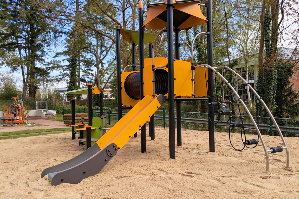 Spielgerät mit Rutsche, Tunnel, Kletterbogen und Spielhaus auf Sandfläche vor Schulgebäude im Hintergrund.