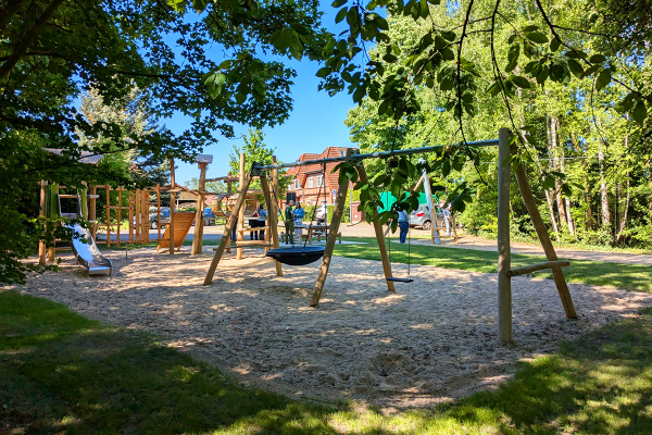 Übersicht über einen naturnahen Spielplatz mit Schaukeln, Rutsche und Klettergerüst, eingebettet in Grünflächen, mit Kindern und Erwachsenen im Hintergrund.