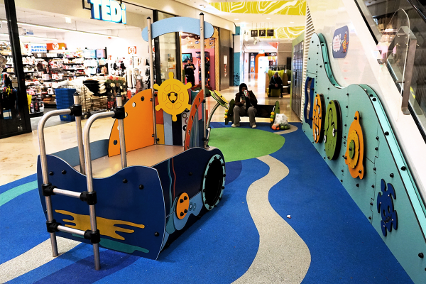 Gesamtansicht eines Indoor-Spielbereichs im Einkaufszentrum mit Spielschiff, taktiler Spielwand „Rheinwelle“ und Fallschutzboden in Blau- und Grüntönen, im Hintergrund Sitzbereiche und Ladenflächen.