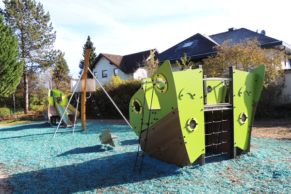 Modernes Schiffswrack-Spielgerät mit Kletterelementen, Rutsche und Schaukel auf blauem Fallschutzbelag, im Hintergrund Wohnhäuser und Bäume.