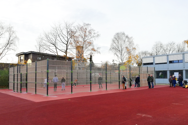 Kleinspielfeld mit rotem Kunststoffbelag und umlaufendem Ballfangzaun auf dem Schulhof der KGS Bad Bevensen, mit barrierefreiem Zugang und zuschauenden Personen.