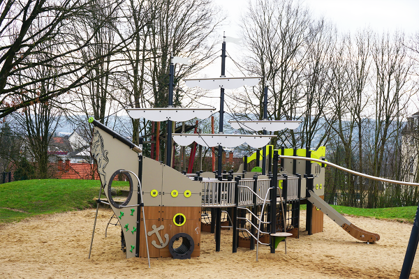 Maritimes Spielschiff mit Rutsche, Netz- und Kletterelementen auf einem Spielplatz mit Sandboden und natürlicher Umgebung