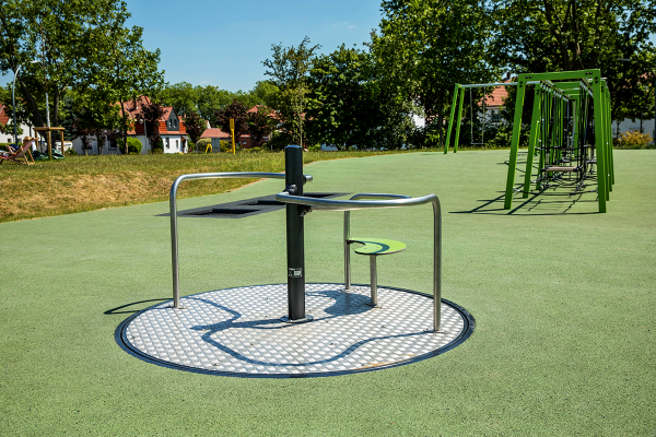 Niedriges, rundes Karussell mit Handlauf und Sitzfläche auf fugenlosem, grünem Fallschutzboden. Es ist barrierefrei zugänglich und für Rollstuhlnutzer geeignet.