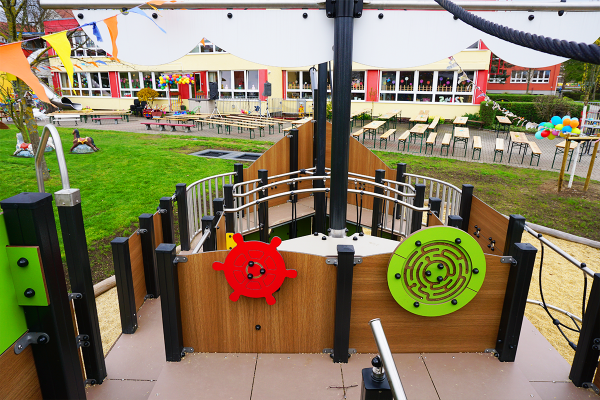 Detailaufnahme des Spielschiffs „Neptun“ mit roten und grünen Spieltafeln in Steuerform, Blick auf die Kita mit festlich dekorierten Sitzgarnituren im Hintergrund.