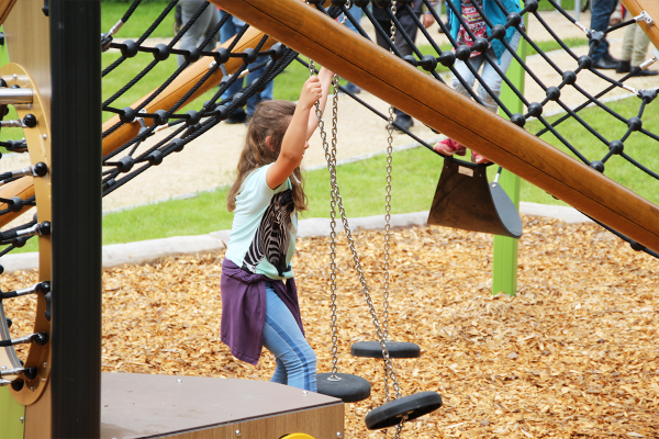 Ein Mädchen balanciert auf Pendelplatten unter einem Spielgerüst mit Kletternetz, während es sich an Ketten festhält.