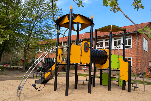 Spielanlage mit Rutsche, Tunnel und Kletterbogen auf Sandfläche vor einem roten Schulgebäude mit weißen Fenstern.