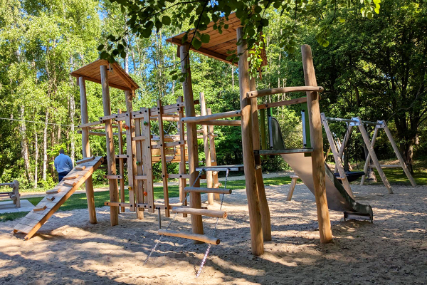 Spielplatz mit großer Kletterstruktur aus Holz, Kletterwänden, Seilaufstieg und Edelstahlrutsche, umgeben von Bäumen und Sandfläche.