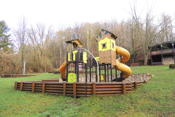 Spielanlage mit zwei Spieltürmen, Röhrenrutsche, Kletterwand und Brücke im Naturpark Kallenbach, eingefasst von einer Holzumrandung und Natursteinmauer, im Hintergrund eine Schutzhütte
