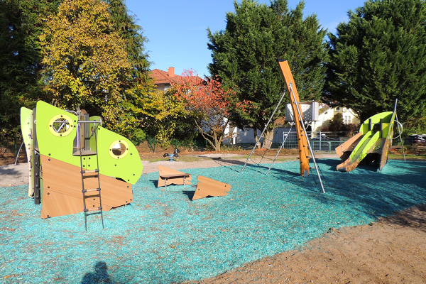 Spielplatz mit grün-braunem Schiffswrack-Spielgerät, Rutsche, Kletterelementen, Schaukel und zwei Sitzbänken auf blauem Fallschutzbelag, umgeben von Bäumen und Wohnhäusern.