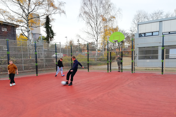 Schüler beim Fußballspiel auf einem barrierearmen Kleinspielfeld mit rotem Kunststoffbelag, umlaufendem Ballfangzaun und integrierter Basketballanlage auf dem Schulhof der KGS Bad Bevensen.