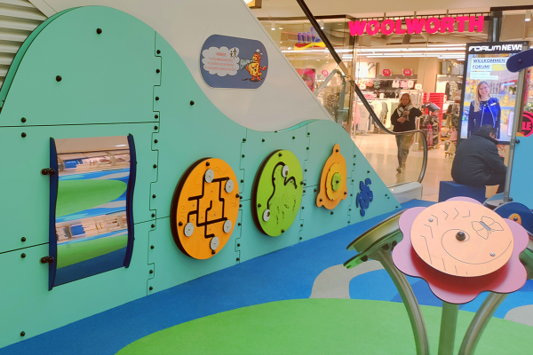 Spielwand mit bunten, drehbaren Spielmodulen in Wellenform, darunter Labyrinth-, Spiegel- und Tastspielelemente, integriert in einen Indoor-Spielbereich im Einkaufszentrum.