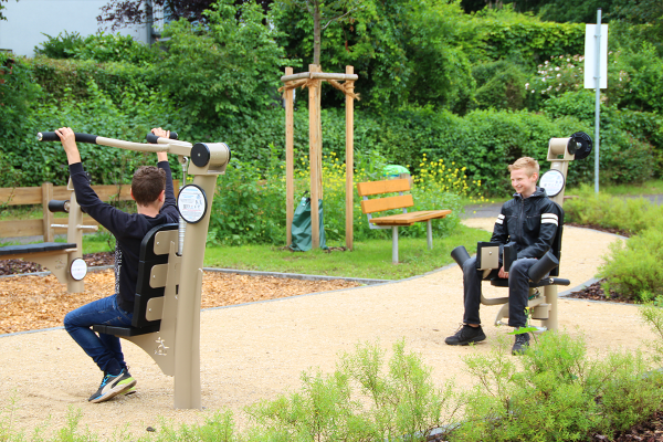 Zwei Jugendliche nutzen Outdoor-Fitnessgeräte auf einem Spielplatz: einer am Schultertrainer, der andere an einer Abduktoren-Maschine.