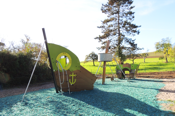 Spielplatz mit thematischem Schiffswrack-Spielgerät und Schaukel auf blauem Fallschutzbelag, in ländlicher Umgebung mit Wiesen und Bäumen.