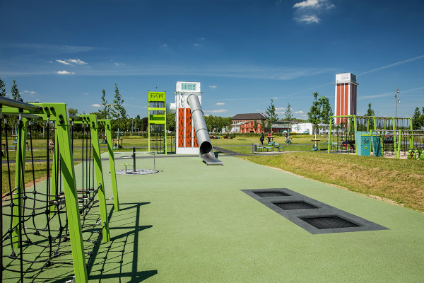 Drei rechteckige Bodentrampoline auf grünem Fallschutzboden, daneben ein barrierefreies Karussell, Kletternetze und ein großer Spielturm mit Edelstahlrutsche im Hintergrund.