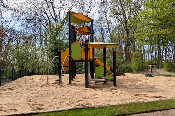 Spielgerät mit Rutsche, Kletterelementen und Plattformen auf einem Sandspielplatz, umgeben von Bäumen.