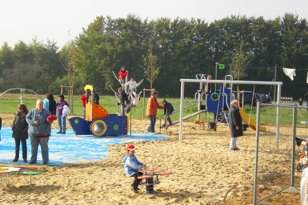 Kinder spielen auf verschiedenen Spielgeräten wie Spielboot, Kletteranlage, Rutsche und Federwippe auf einem Spielplatz mit Sand- und Wasserfläche, begleitet von Erwachsenen