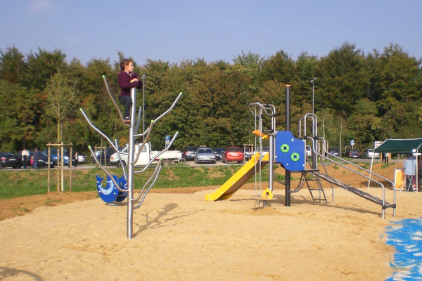 Spielplatzszene mit Kind auf Klettergerät im Vordergrund und blauem Spielturm mit Rutsche, Schaukel und Kletterrampe im Hintergrund