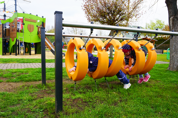 Zwei Kinder krabbeln durch einen hängenden Reifenparcours aus orangefarbenen Kunststoffringen auf einem Kita-Spielplatz mit Spielschiff im Hintergrund.