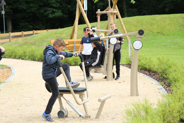 Ein Junge trainiert an einem Outdoor-Rudergerät, im Hintergrund nutzen weitere Kinder Fitnessgeräte in einem Park.