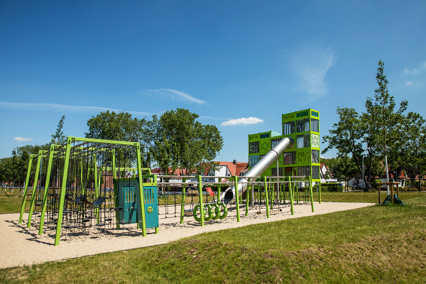 Grünes Klettergerüst mit Seilen, Schaukeln und einem hohen Kletterturm mit Edelstahlrutsche auf einer Sandfläche, eingebettet in eine parkartige Umgebung mit Bäumen und Wohnbebauung im Hintergrund.